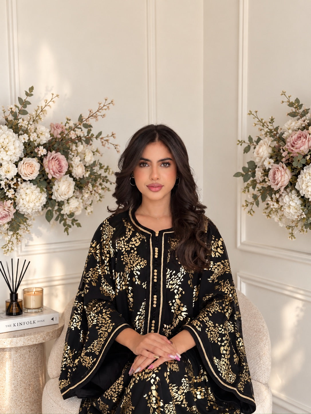 Model seated in black Gandoura Raya with a gold-toned floral pattern and wide sleeves